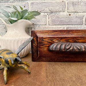 May include: A dark brown, rectangular wooden box with a carved, decorative handle. The box has a rich, reddish-brown stain and a detailed, floral-like design on the handle. A decorative bee and a white pitcher with greenery are also in the image.