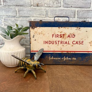 May include: A vintage metal first aid industrial case with a blue and white design, featuring the text "FIRST AID INDUSTRIAL CASE" in red. A decorative bee figurine is in the foreground, and a white vase with greenery is on the left.