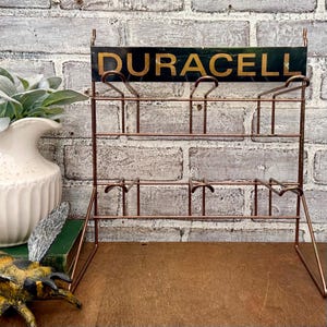 May include: A vintage copper-colored wire display rack with the word "DURACELL" in gold lettering on a dark green sign. The rack has multiple tiers for holding items and is set against a brick wall.