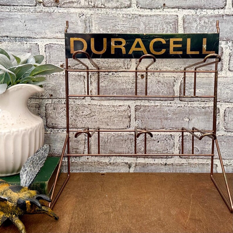 May include: A vintage copper-colored wire display rack with the word "DURACELL" in gold lettering on a dark green sign. The rack has multiple tiers for holding items and is set against a brick wall.