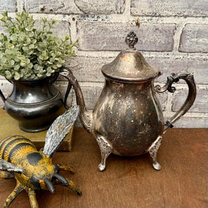 May include: Antique silver teapot with ornate details, including a curved handle and decorative feet. The teapot has a lid with a finial and a spout. A decorative bee figurine is in the foreground, and a small vase with greenery is in the background.