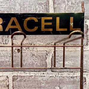 May include: A vintage metal sign with the word "RACELL" in gold lettering on a dark green background. The sign has a copper-colored metal frame with hooks and a grid-like structure, mounted on a textured brick wall.