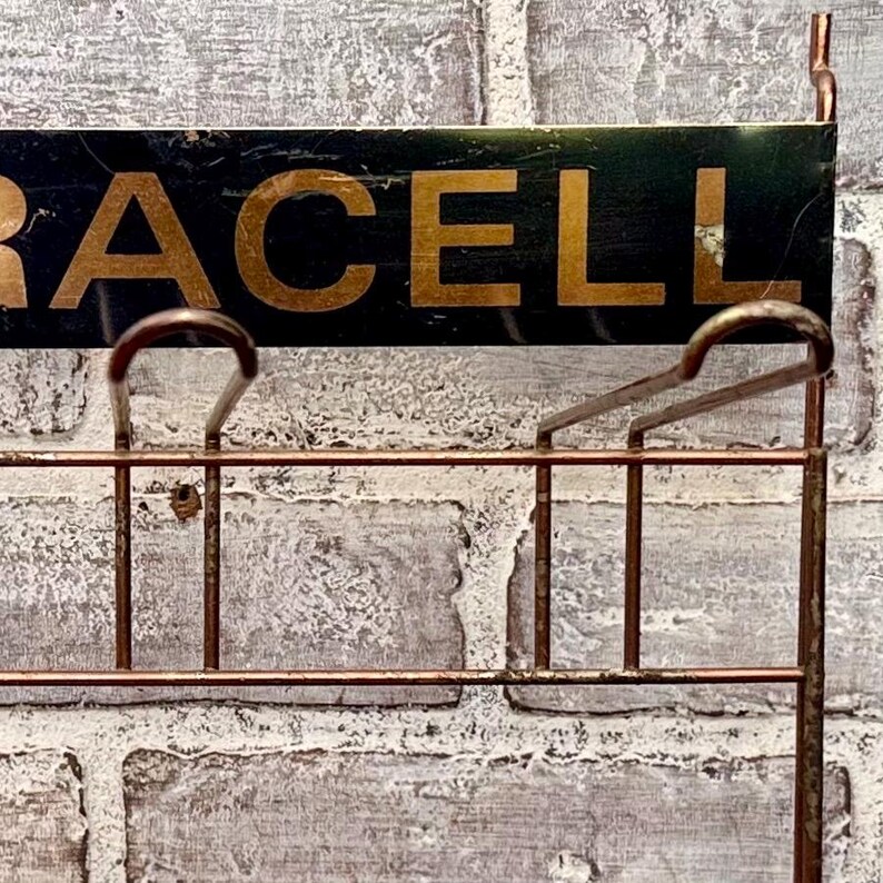 May include: A vintage metal sign with the word "RACELL" in gold lettering on a dark green background. The sign has a copper-colored metal frame with hooks and a grid-like structure, mounted on a textured brick wall.