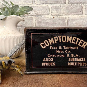 May include: A black rectangular metal sign with orange and cream text reading COMPTOMETER, FELT & TARRANT MFG. CO., CHICAGO, U.S.A., ADDS, SUBTRACTS, DIVIDES, MULTIPLIES. The sign features a thin orange border and shows signs of wear on the edges.