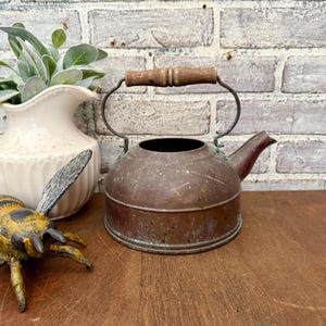 May include: A vintage copper kettle with a wooden handle and spout. The kettle has a rustic, aged appearance, with visible wear and patina. The handle is made of light brown wood. The kettle is sitting on a wooden surface.