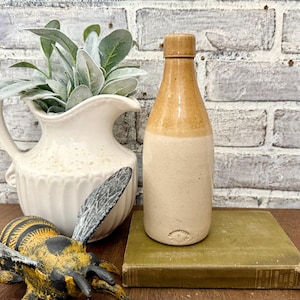 May include: A vintage stoneware bottle with a tan and cream color scheme. The bottle has a rounded body and a narrow neck with a stopper. It is resting on a green book. A white pitcher with greenery and a decorative bee are also in the image.