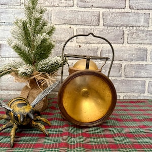 May include: An antique brass lantern with a round, golden reflector and a dark brown wooden frame. The lantern has a metal handle and sits on a red and green plaid surface. A decorative bee and a small Christmas tree are also in the image.