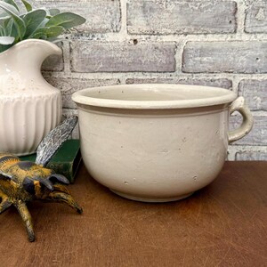 May include: A cream-coloured, round ceramic bowl with a handle. The bowl has a wide rim and a slightly textured surface. The background includes a white jug, a decorative bee, and a brick wall.