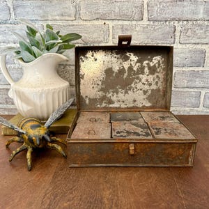 May include: An open, antique metal box with six small compartments inside. The box has a rusty, distressed finish. A decorative bee figurine and a white pitcher with greenery are next to the box.