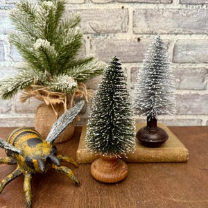 May include: A festive display featuring miniature Christmas trees, one green and one silver, on a wooden surface. A rustic bee sculpture and a small evergreen plant in a burlap pot are also present.