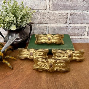 May include: Five gold-colored decorative bow-shaped ornaments with intricate leaf and bead detailing. The ornaments are arranged on a wooden surface, with a green book in the background. The bows have a central oval accent.