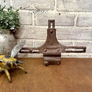 Peut inclure: Un support en métal marron rouillé, de forme symétrique. Il présente une partie centrale verticale avec une ouverture rectangulaire et des bras horizontaux. L'objet a un aspect vieilli. En arrière-plan, un mur de briques et une surface en bois.