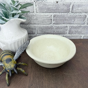 May include: A cream-coloured ceramic bowl with a slightly textured surface. The bowl is round with a wide rim and sits on a dark wooden surface. A decorative bee figurine and a white vase are also in the image.