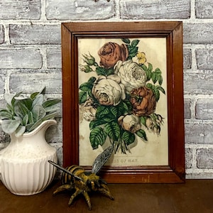 May include: A framed floral print featuring a bouquet of roses in shades of white, peach, and brown, with green leaves. The print is in a dark brown wooden frame. A decorative bee figurine and a white vase with greenery are in the foreground.