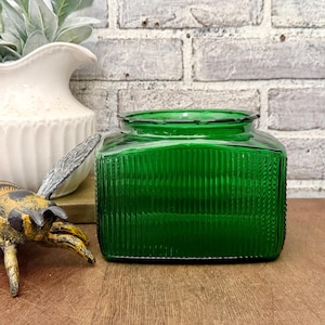 May include: A square, emerald green glass jar with vertical ribbed detailing. The jar has a slightly scalloped rim and sits on a wooden surface. A white vase and a decorative bee figurine are partially visible in the background.