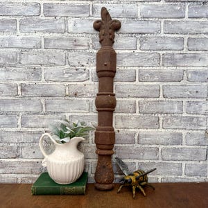 May include: A tall, weathered, brown metal architectural element with a fleur-de-lis top. The column has a series of decorative rings and a hexagonal midsection. A white pitcher with faux greenery, a green book, and a bee figurine are in the foreground.