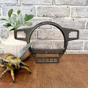 May include: A vintage metal Zenith gauge bezel with a circular opening and rectangular side mounts. The word "ZENITH" is embossed across the bottom. The bezel is displayed with a decorative bee figurine and a white vase with greenery.