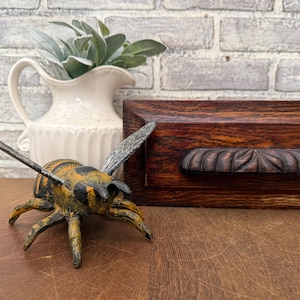 May include: A decorative metal bee figurine with yellow and black stripes, wings, and legs. A white pitcher with green leaves and a dark wooden box with a carved handle are in the background.