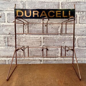 May include: A vintage copper-toned metal display rack with the word "DURACELL" in gold lettering on a black sign. The rack has two tiers for holding items and is set against a brick wall.
