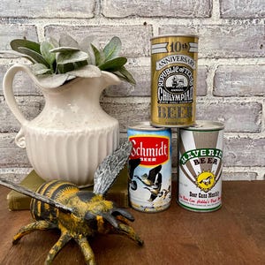 May include: Three vintage beer cans with different designs. The cans are on a wooden surface with a white pitcher and a bee figurine in the foreground.
