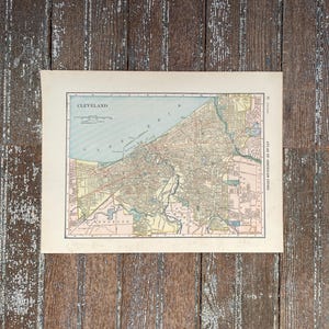 May include: A vintage map of Cleveland, Ohio, printed in black and white with colored accents. The map shows the city's streets, parks, and waterways. The title "CLEVELAND" is printed in the upper left corner, and "ATLAS OF AMERICAN CITIES" is printed in the upper right corner.