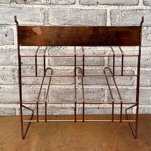 May include: A copper-colored metal magazine rack with a rectangular top and a grid-like design. The rack has a series of vertical and horizontal bars, with curved supports. The rack is designed to stand upright and is suitable for organizing reading materials.