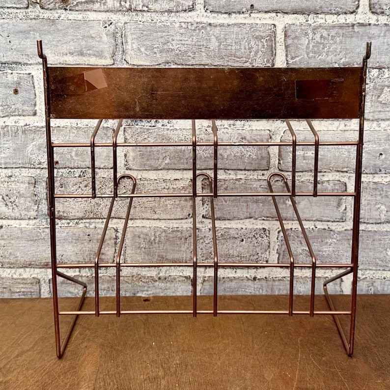 May include: A copper-colored metal magazine rack with a rectangular top and a grid-like design. The rack has a series of vertical and horizontal bars, with curved supports. The rack is designed to stand upright and is suitable for organizing reading materials.