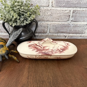 以下が含まれることがあります： 楕円形のアンティークセラミック皿、蓋付き。 皿はオフホワイトで、マルーンの花柄と風景のデザインが施されています。 蓋には取っ手があり、皿は木製の表面にあります。 装飾的な金属の花瓶と造花が背景にあります。