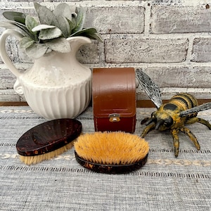 May include: Two brown and black handled brushes with natural bristles, a brown leather case, and a yellow and black bee figurine on a gray and white patterned surface.