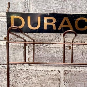May include: A vintage-style wall-mounted rack with the word "DURAC" in gold lettering on a black background. The rack is made of copper-colored metal and is mounted on a weathered brick wall.