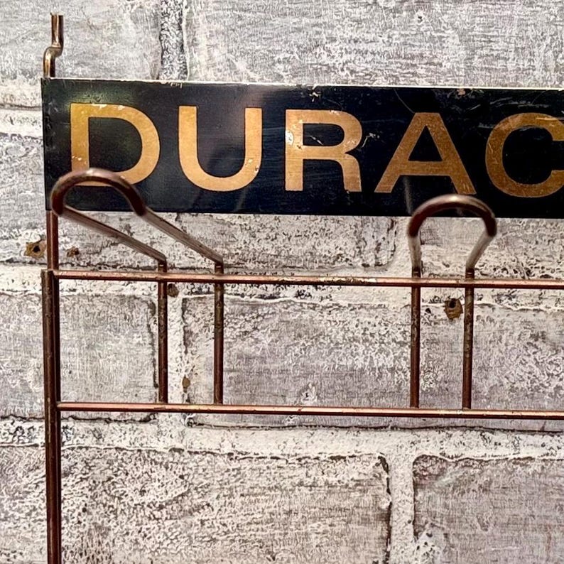 May include: A vintage-style wall-mounted rack with the word "DURAC" in gold lettering on a black background. The rack is made of copper-colored metal and is mounted on a weathered brick wall.