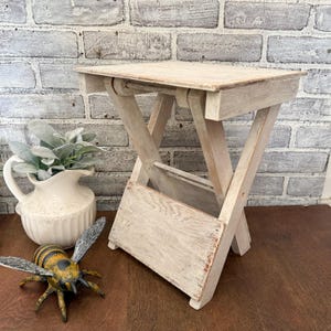 Peut inclure: Petite table d'appoint pliante en bois avec une finition blanc cassé vieillie. La table a un plateau carré et un piètement en X. Une figurine d'abeille décorative et un pichet blanc avec de la verdure sont également dans l'image.