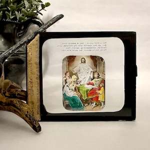 May include: A framed religious print featuring a depiction of Jesus with figures in a room. The print has a black frame and includes text at the top. The image is a vintage piece.