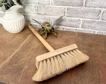May include: A wooden-handled broom with natural bristles. The broom is lying on a brown wooden surface. A yellow and black bee figurine is in the background.