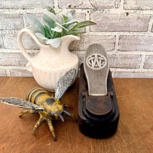 May include: A cast iron bee figurine with yellow and black stripes, a cream-colored ceramic pitcher containing green foliage, and a black metal object featuring a silver oval plate embossed with the letters CWG in a circular frame.
