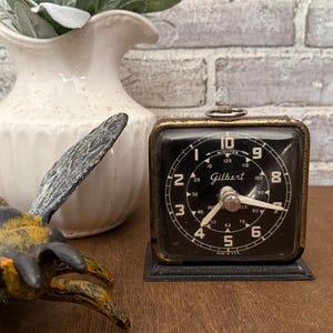 May include: A vintage square Gilbert interval timer with a black face, white numerals, and a metallic gold-toned frame. The dial features markings for minutes up to 120, with white hands and a small metal ring on top, resting on a black rectangular base.