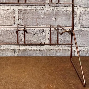 May include: A copper-colored metal display rack with multiple shelves and hooks. The rack is designed to stand upright and has a simple, minimalist design. The background is a textured, light-colored brick wall.