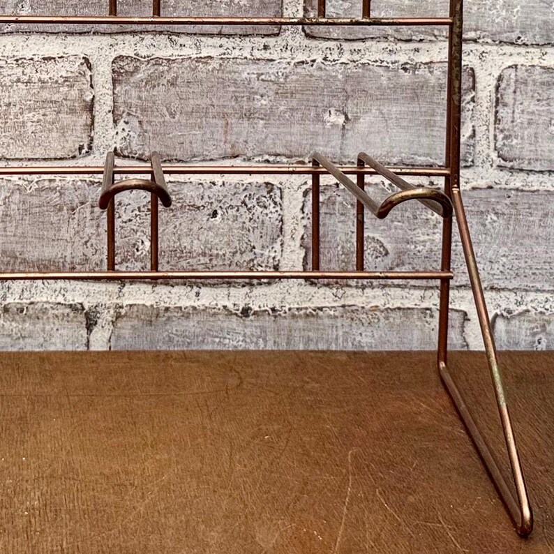 May include: A copper-colored metal display rack with multiple shelves and hooks. The rack is designed to stand upright and has a simple, minimalist design. The background is a textured, light-colored brick wall.