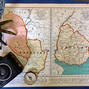 May include: A vintage map of Paraguay and Uruguay, with a compass, a toy bee, and a vintage camera on top of it. The map is printed on paper with a blue border and has a light brown background. The map shows the countries' borders, rivers, and cities.