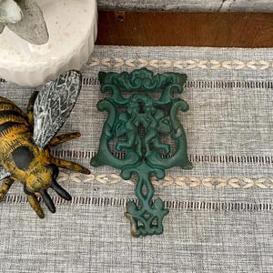 May include: A green cast iron trivet with a decorative design of two cherubs holding a heart. The trivet has a scalloped edge and a floral design.