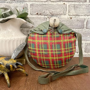 May include: A vintage green canvas canteen with a plaid pattern and a metal strap. The canteen has a metal lid and a metal chain attached to the lid.