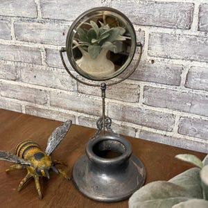 May include: A vintage-style, silver-toned vanity mirror with a round, magnifying glass. The mirror is mounted on a decorative stand with a wide base. A decorative bee figurine is on the left. The mirror reflects a white vase with green leaves.