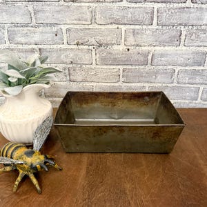 May include: A rectangular, aged metal container with a slightly trapezoidal shape. The container has a weathered, rustic appearance, with visible signs of age and use. The background features a white brick wall and a wooden surface.