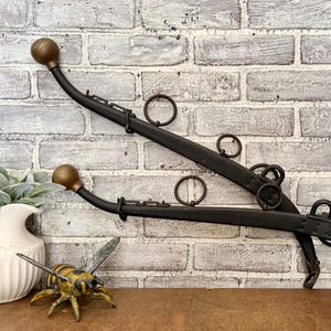 May include: A black metal harness yoke featuring two curved arms with brass-colored spherical finials at the ends and several attached metal rings. The object is positioned horizontally, showcasing its dark, textured finish and structural design.