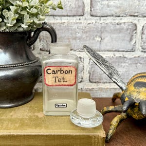May include: A vintage clear glass bottle labelled "Carbon Tet." and "Upjohn." A clear glass stopper sits beside it. The bottle rests on a green book. The background includes a decorative bee.