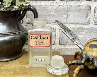 May include: A vintage clear glass bottle with a label that reads "Carbon Tet." and "Upjohn." The bottle has a clear glass stopper next to it. The bottle is on a green book.