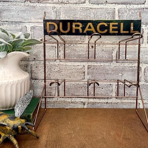 May include: A vintage copper-toned metal display rack with the word "DURACELL" in gold lettering on a dark green background. The rack has multiple tiers for displaying items.