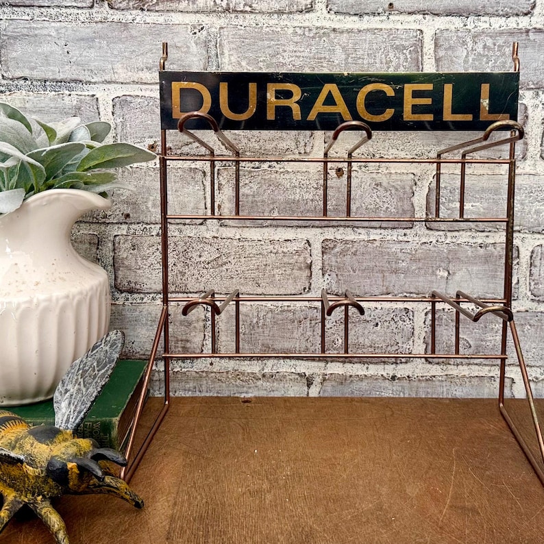 May include: A vintage copper-toned metal display rack with the word "DURACELL" in gold lettering on a dark green background. The rack has multiple tiers for displaying items.