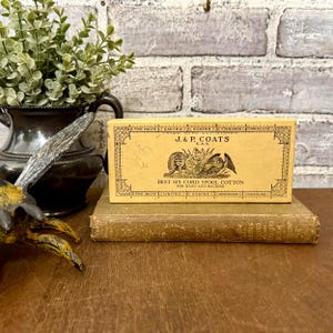 May include: A vintage box of J. & P. Coats Best Six Cord Spool Cotton, with text and illustrations, rests on a worn book. The box is cream-colored with black text and a decorative border. A small, green plant in a dark vase is in the background.