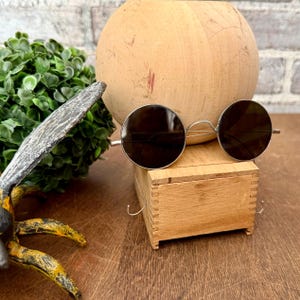 May include: Round sunglasses with dark lenses and a silver frame rest on a small wooden box. A large, round, light-colored wooden object is in the background. A decorative bee is in the foreground.